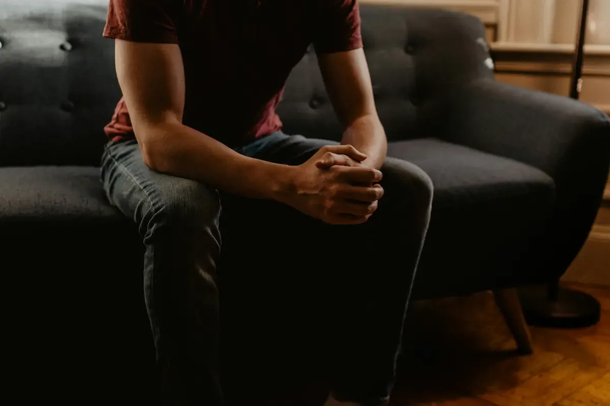 Close-up of a man sitting on the edge of a gray couch with hands clasped and forearms resting on his knees, his face out of frame, conveying a pensive mood.