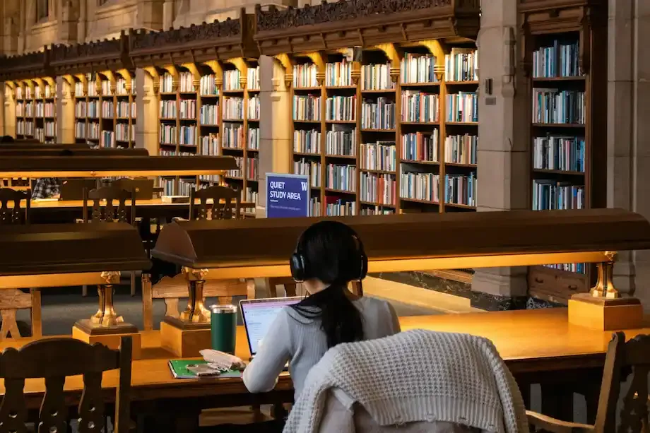 Student wearing headphones studies on a laptop at a wooden desk in a quiet university library lined with bookshelves